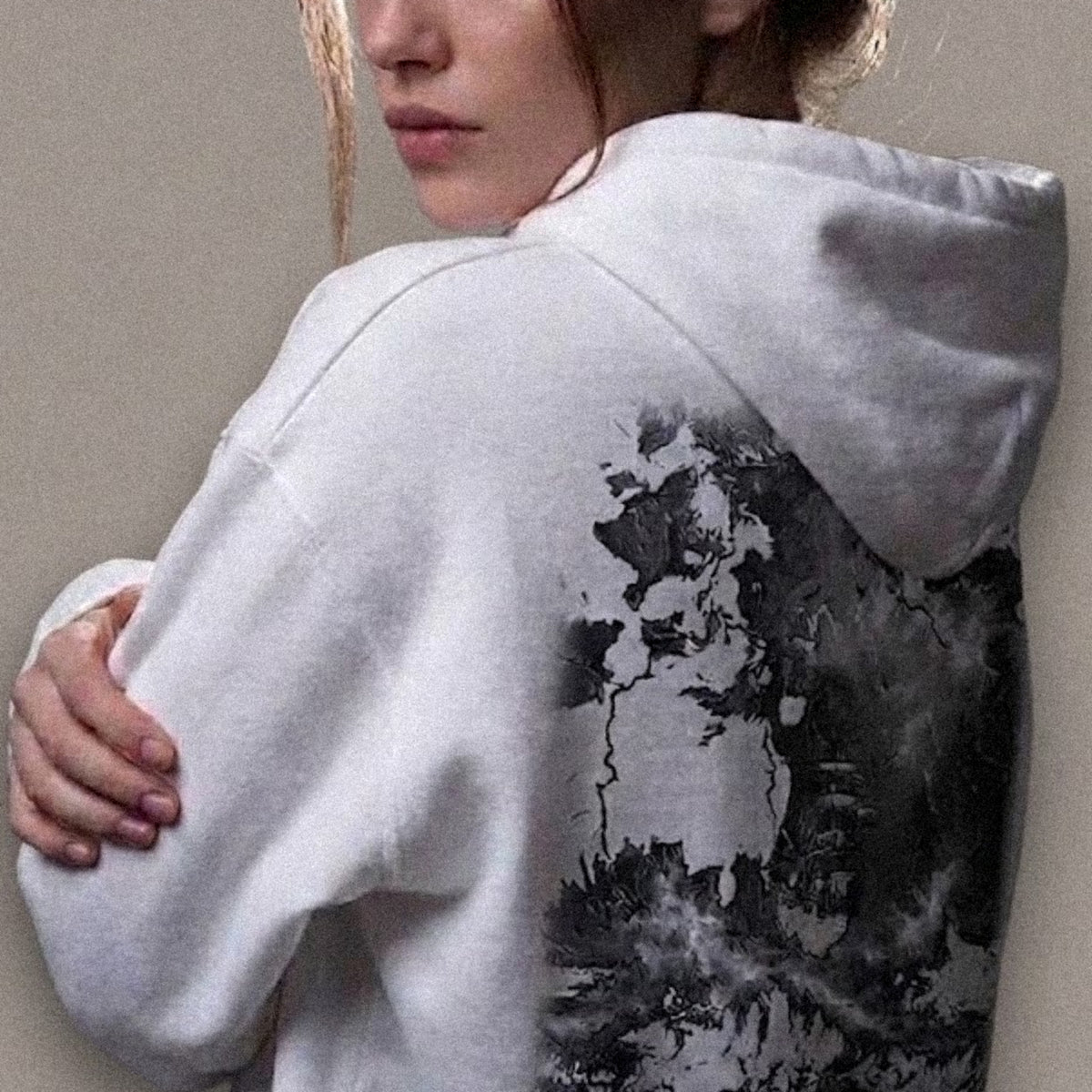 A woman wearing the relaxed-fit hoodie in white, seen from behind, showing the ABYSS design printed across the back: a tall-masted ship heeled over against rising swell and closing storm, in monochrome greys.